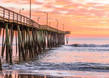 Wooden pier extending over gentle ocean waves at sunrise, with a colorful pink and orange sky.