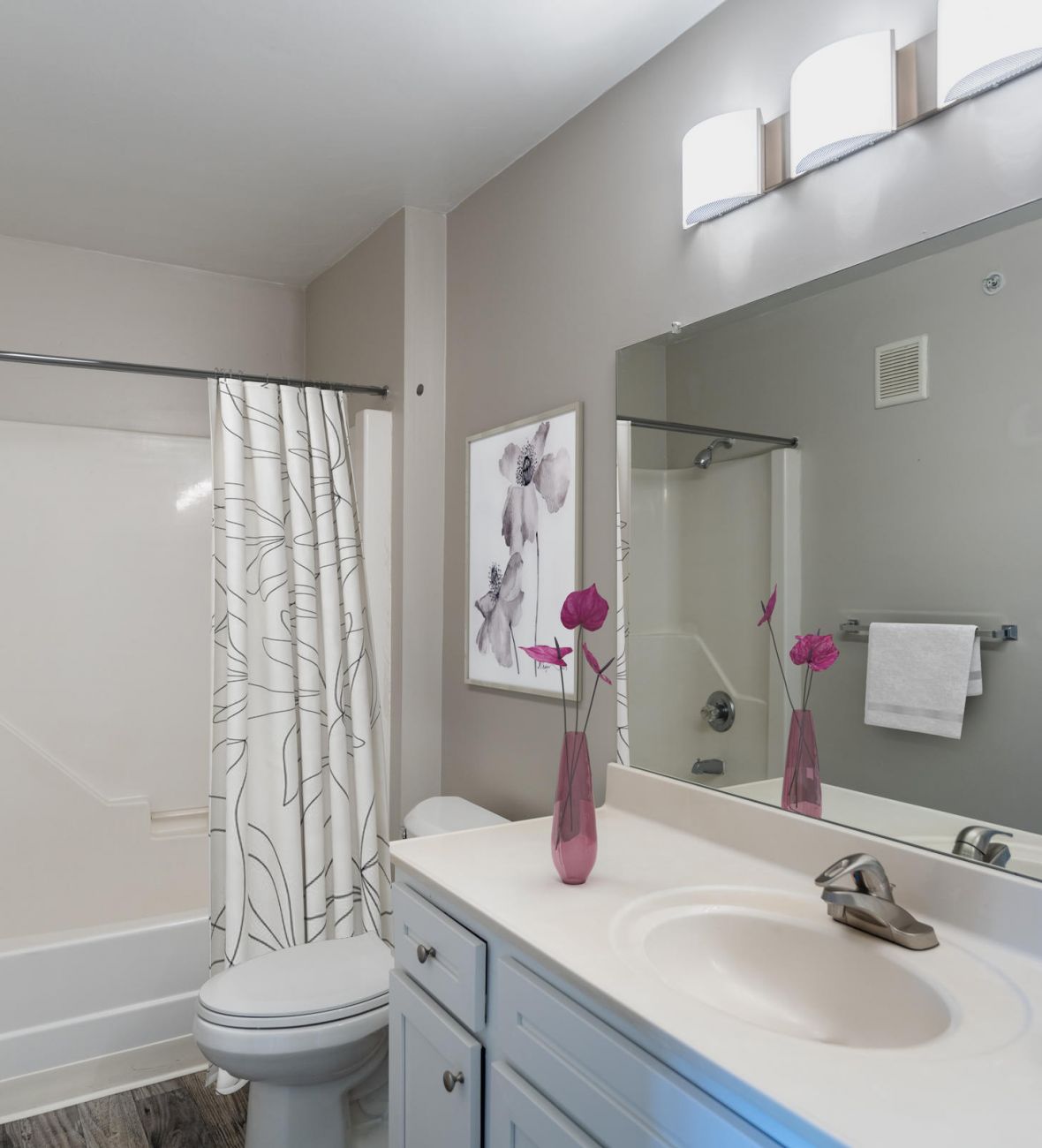 Attain on 5th apartment homes with Modern bathroom with white vanity, large mirror, shower curtain, and purple flowers in a vase on the counter.