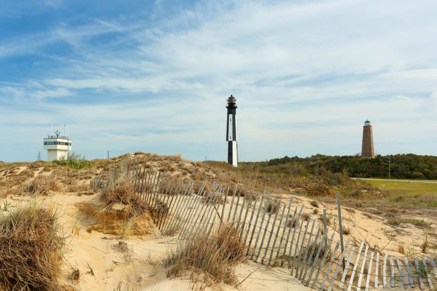 A sandy dune with a weathered fence, two lighthouses, and a modern building under a blue sky.