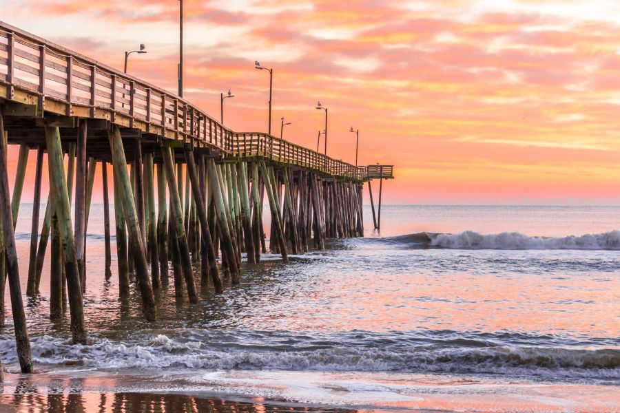 Wooden pier extending over gentle ocean waves at sunrise, with a colorful pink and orange sky.