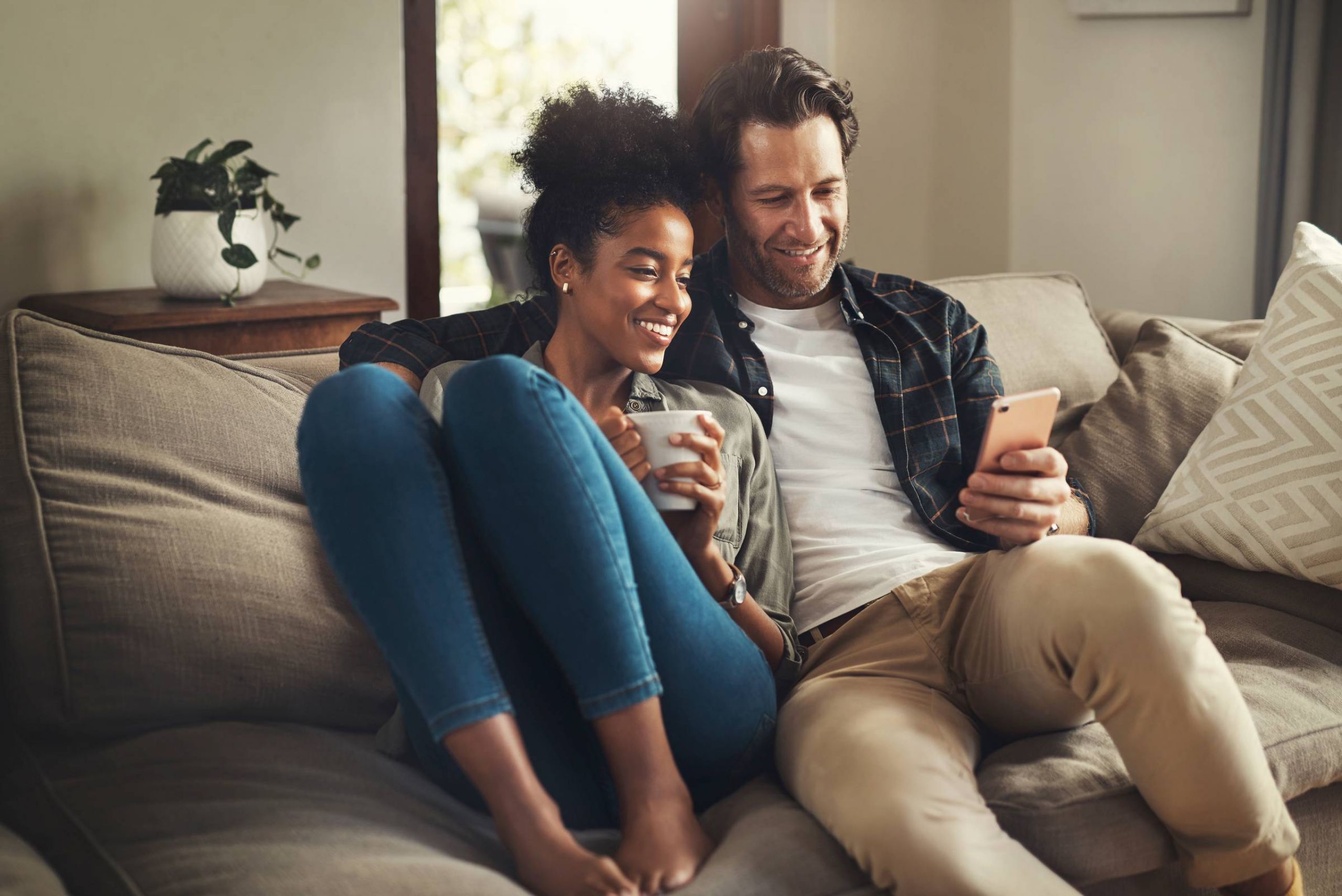 Attain on 5th apartment homes with Smiling couple sits on a couch together, looking at a smartphone and holding a mug.